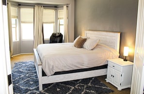 Un dormitorio con cama, mesita de noche, lámpara, silla y ventana con cortinas.