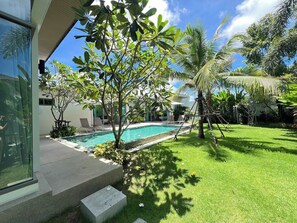 A swimming pool surrounded by lush greenery, a clear sky, and a modern building with large windows. - Serenity Villa With Private Pool, Phuket (Cherngtalay)