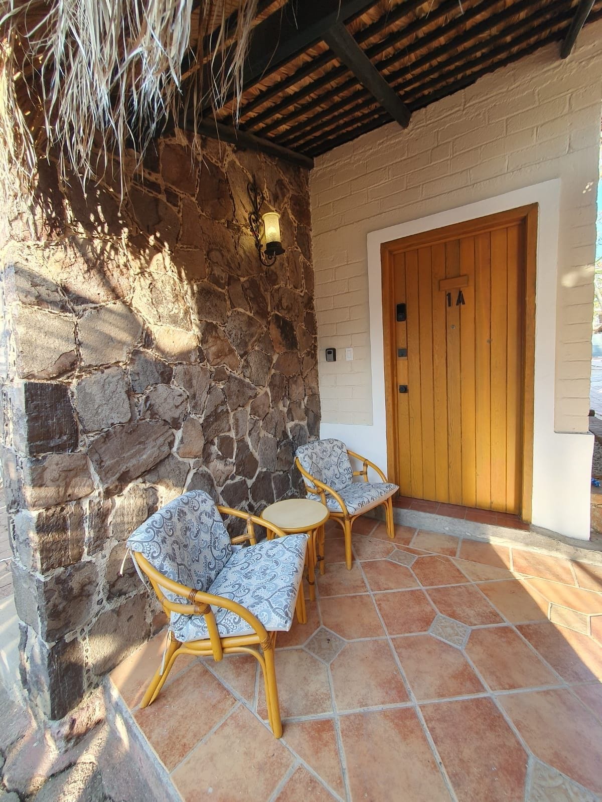 A stone wall with a wooden door, two wicker chairs with patterned cushions, and a tiled patio.