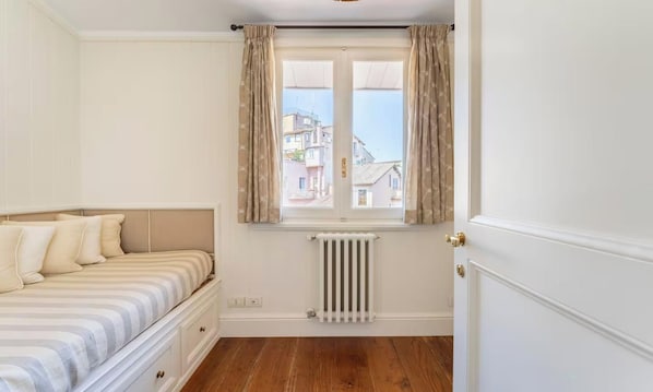 Et soveværelse med seng, radiator, et vindue med gardiner og udsigt til bygninger.