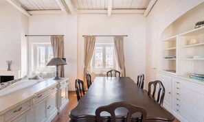 Een eetkamer met een lange tafel, stoelen, een lamp en een boekenplank.