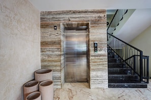 En elevator med metalfinish, væg med marmor-mønster og trappe med sorte gelændere.