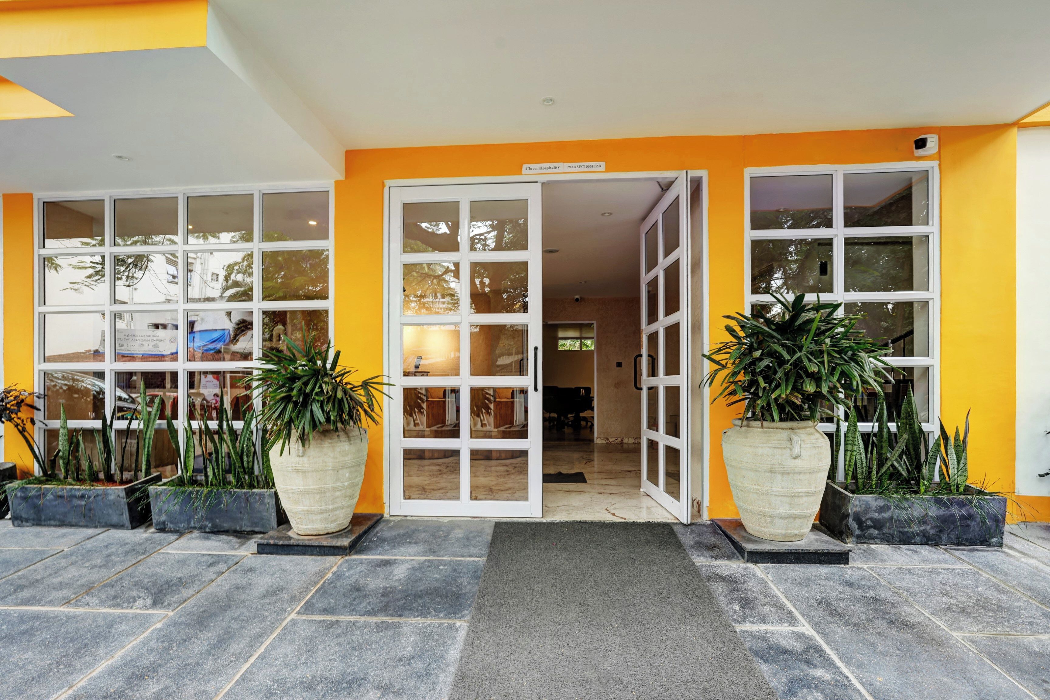 A hotel entrance with a yellow facade, large potted plants, and a tiled entrance area.