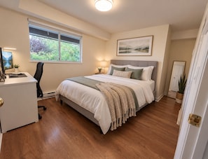 A bedroom with a bed, desk, chair, and a window with a view of greenery. - Large 1 Bedroom private Suite (Coquitlam)