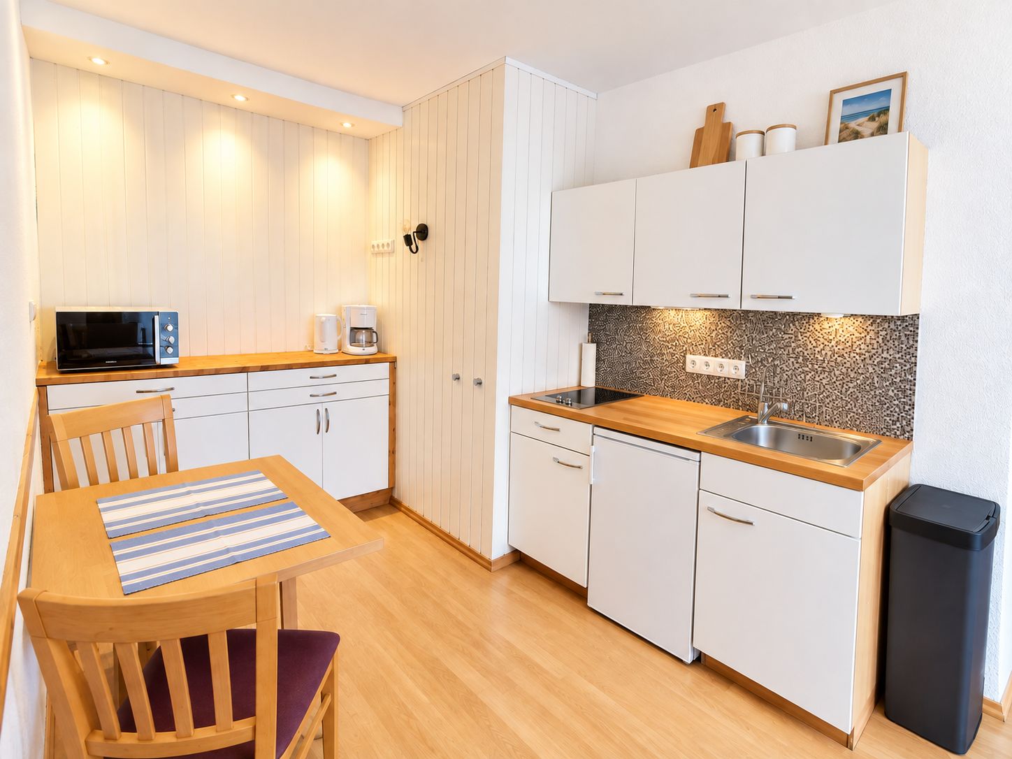 Una cocina compacta con armarios blancos, microondas, cafetera y lavamanos.