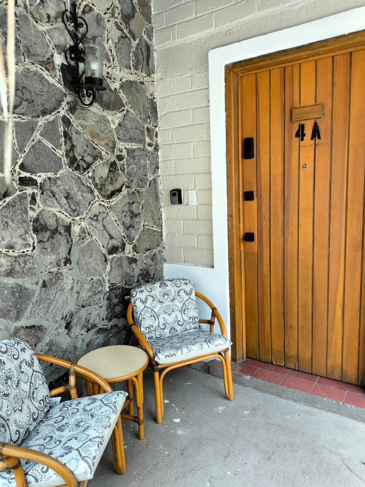 Un muro de piedra con un accesorio de luz metálico, una puerta de madera con un número y dos sillas de mimbre con cojines estampados.