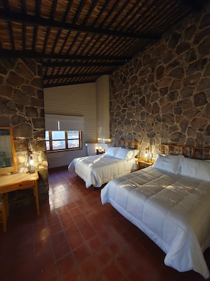 A room with two beds, stone walls, a wooden ceiling, a window, and a small table with a lamp. - Sandy Shores 4A y 4B (La Paz)