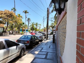 A street lined with parked cars and palm trees, with a building featuring a brick facade and a decorative fence.