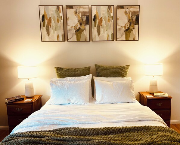 A neatly made bed with white linens and a green knitted throw, flanked by two bedside tables with lamps, and a wall with three abstract paintings.