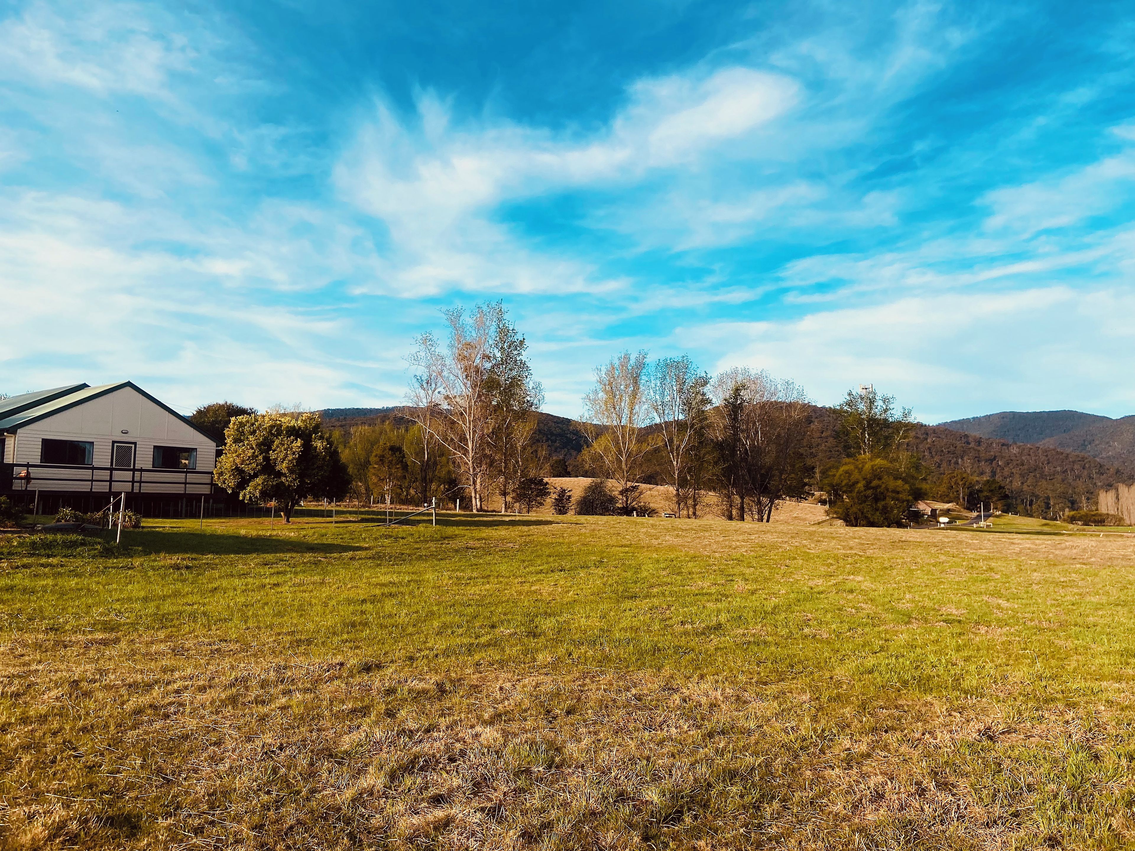 Paesaggio rurale con un ampio campo aperto, un edificio bianco con tetto verde e una collina boscosa sullo sfondo.