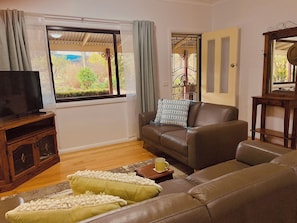A living room with a brown sofa, a television on a wooden stand, a window with curtains, and a view of greenery outside. - The Plonk Shop Cottage - Farm Stay & River Access (Towamba)