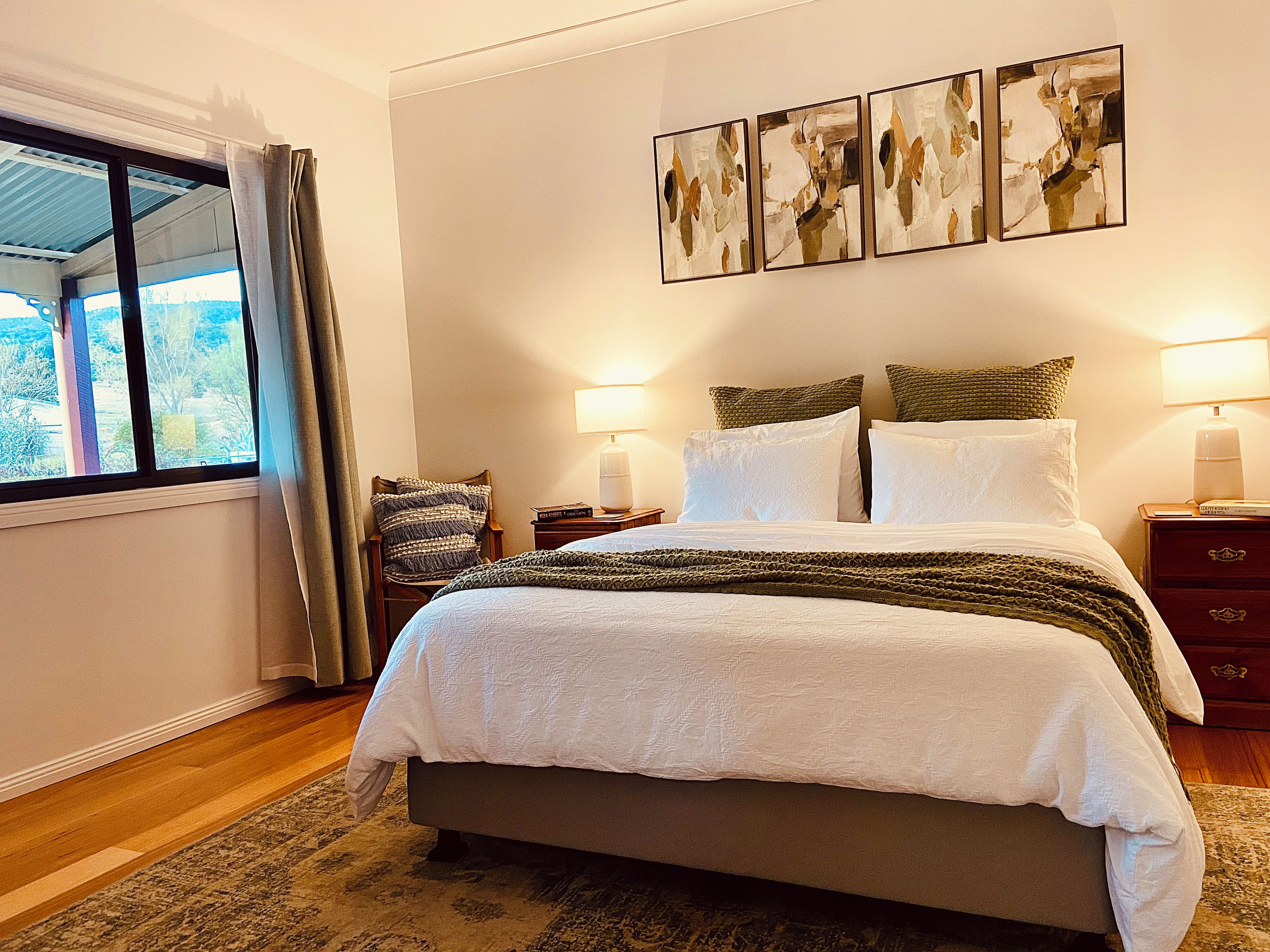 A bedroom with a large bed, a window with curtains, a chair, a bedside table with a lamp, and a wall with three framed artworks.