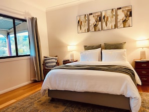 A bedroom with a large bed, a window with curtains, a chair, a bedside table with a lamp, and a wall with three framed artworks. - The Plonk Shop Cottage - Farm Stay & River Access (Towamba)