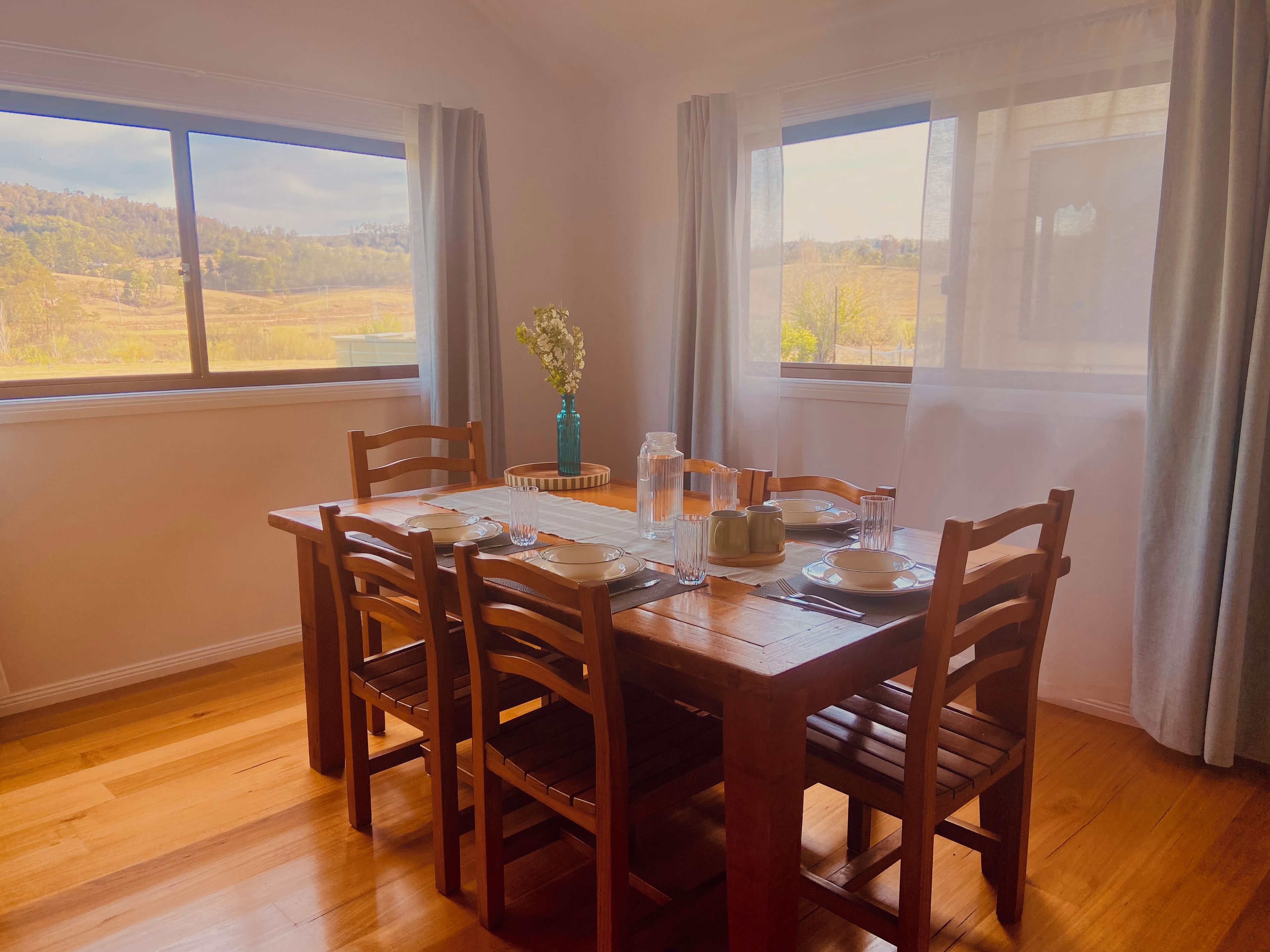 Comedor con una mesa de madera dispuesta para cuatro personas, rodeada de sillas, y vistas a un campo a través de amplias ventanas.