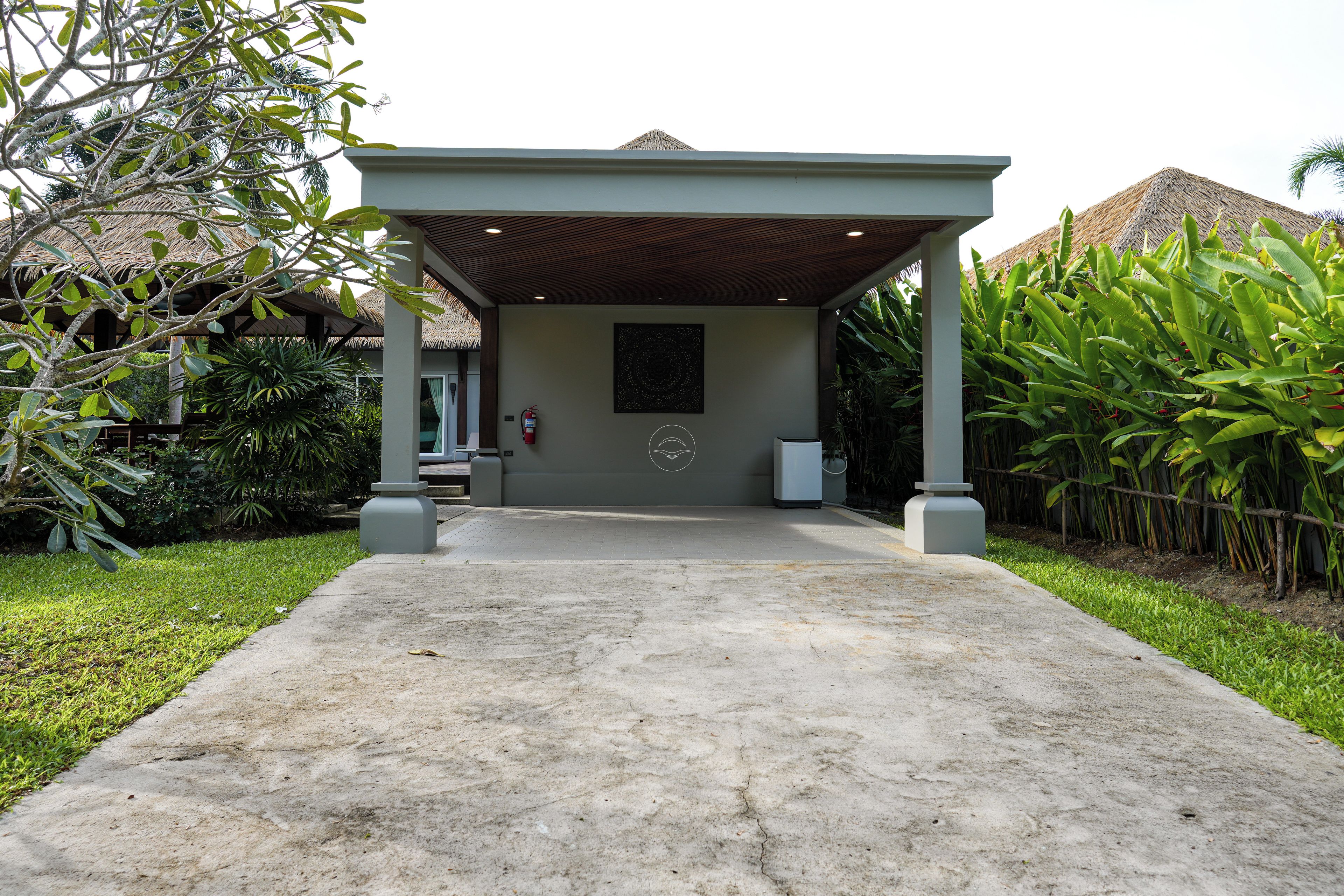 A covered entrance to a building with a concrete pathway leading up to it, surrounded by greenery and a thatched roof structure in the background.