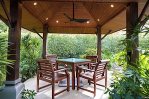 Een houten prieel met een tafel en stoelen, omringd door weelderige begroeiing.
