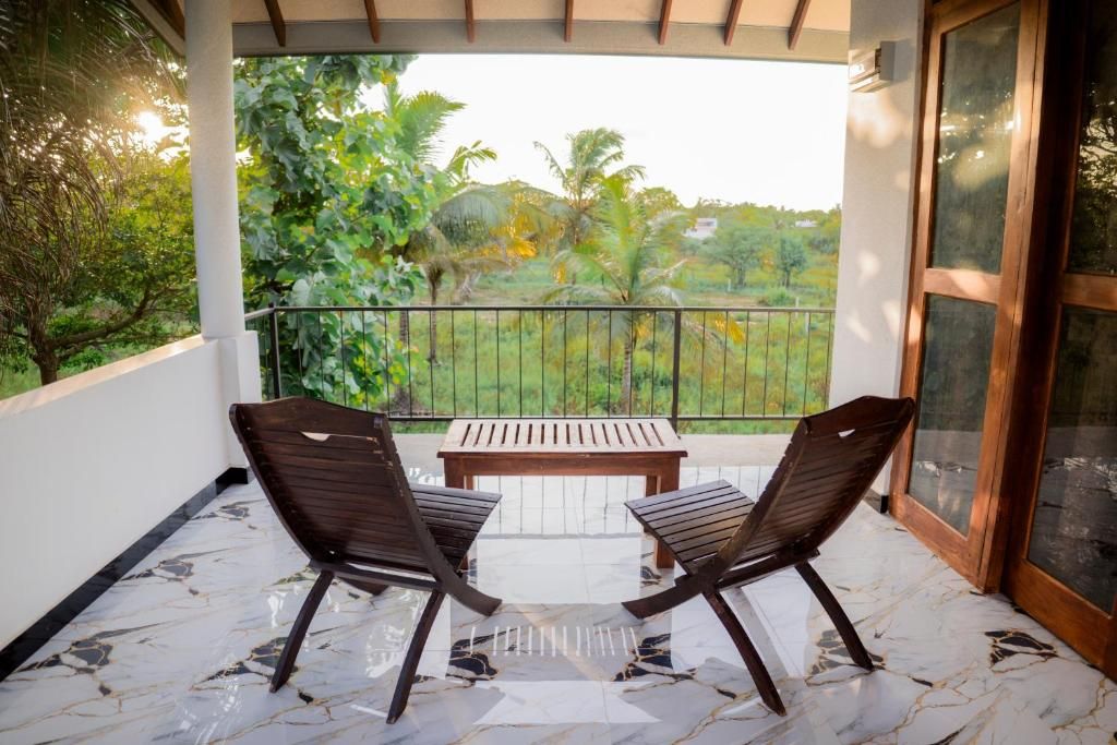 A balcony with two wooden chairs and a table, overlooking a garden with trees and a fence.