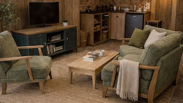 A cozy living room with a television, green armchairs, a coffee table, and a kitchen area in the background.
