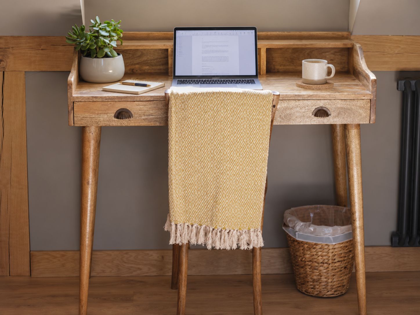 A wooden desk with a laptop, a potted plant, a notebook, a mug, and a trash bin.