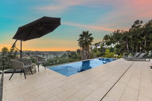 Una piscina infinita al terrat amb vista a la ciutat, cadiretes i una ombrel·la.
