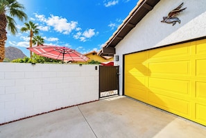 A garage with a yellow door, a white brick wall, and a red umbrella. - Heated Pool • Mountain Views • Fire Pit + Grill (La Quinta)