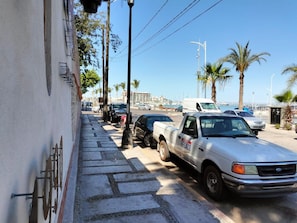 Eine Straße mit geparkten Fahrzeugen, darunter ein weißer Pritschenwagen mit dem Schriftzug 'FESACO' und ein weißer Lieferwagen. Es sind Palmen und ein klarer Himmel zu sehen.