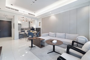 A modern living room with a sectional sofa, a coffee table, and a dining area with chairs and a kitchen in the background. - 1BHK, Burj Khalifa View Business Bay (Dubai)