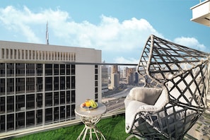 A modern balcony with a glass partition, a wicker chair, a small round table with fruit and a glass, and a cityscape view. - 1BHK, Burj Khalifa View Business Bay (Dubai)