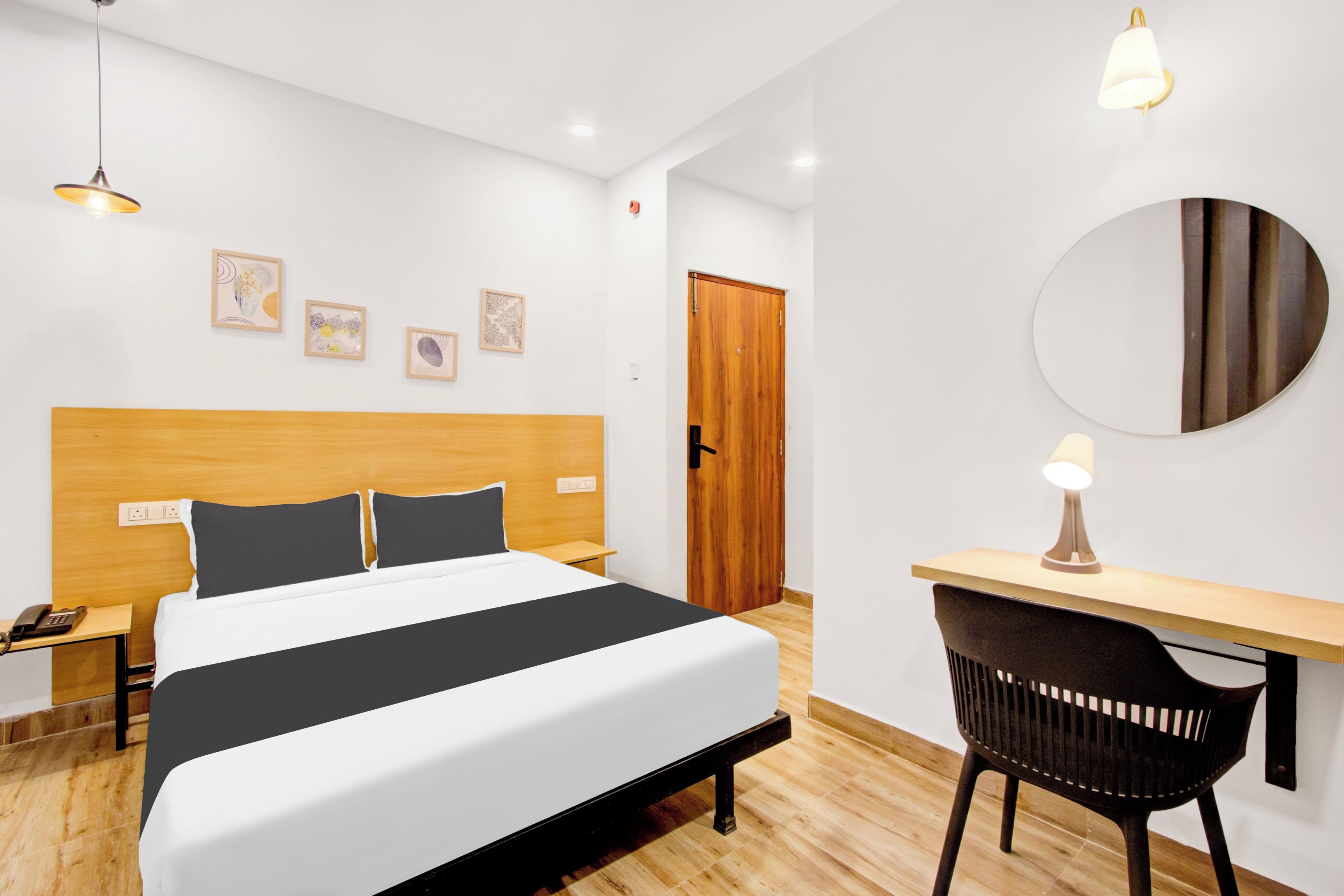 Quarto de hotel moderno com cama de cabeceira em madeira, uma escrivaninha com cadeira, um espelho redondo e três quadros nas paredes.