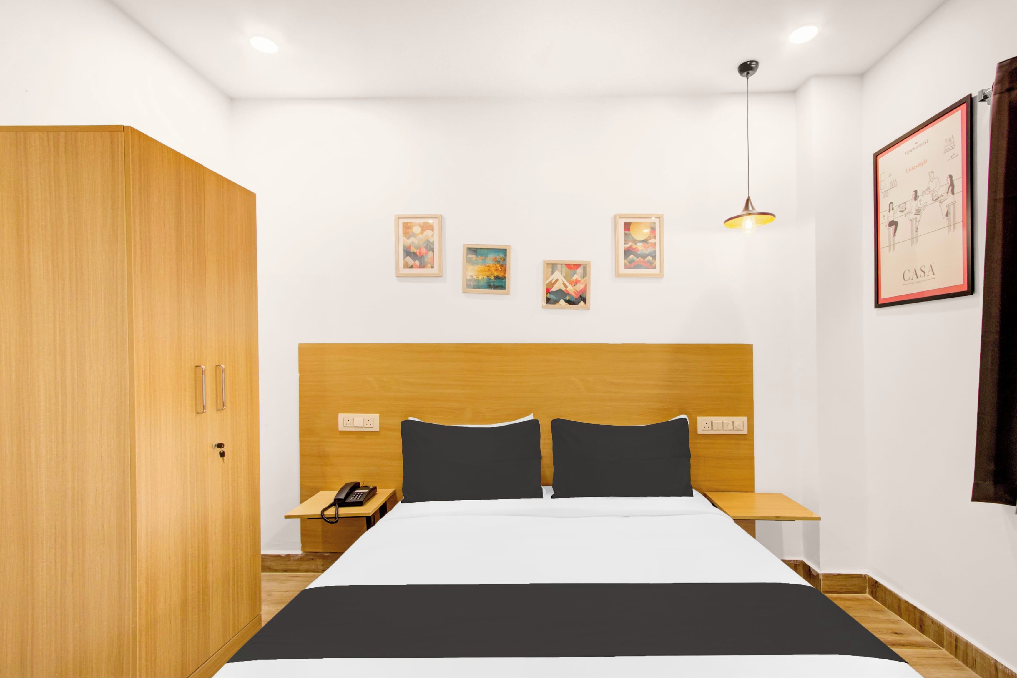 A modern hotel room with a wooden headboard, a bed with black and white bedding, a bedside table with a phone, and a wall-mounted light fixture.