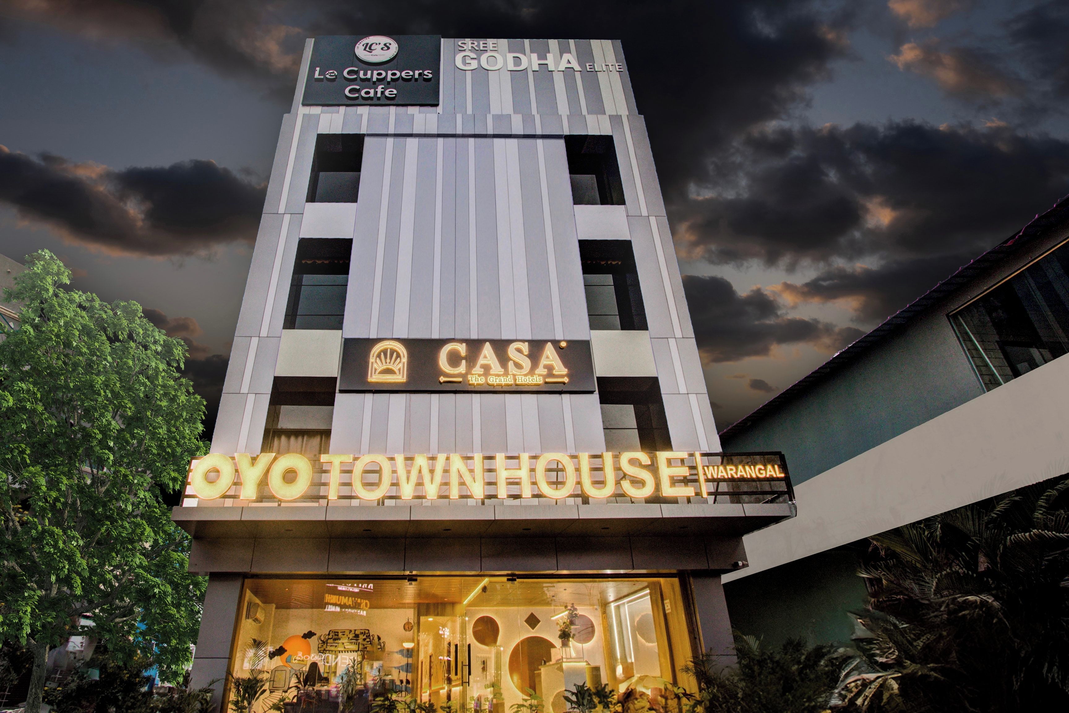 Un edificio de varios pisos con letreros que indican 'Le Cuppers Cafe', 'Sree Godha Elite', 'CASA The Grand Hotels' y 'Oyo Town House'.