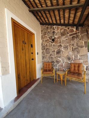 Un muro de piedra con una puerta de madera que dice 'GA' y dos sillas de mimbre con cojines.