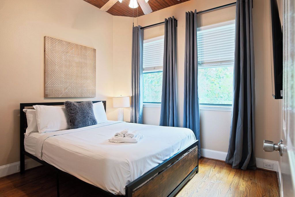 Une chambre à coucher avec un lit, une œuvre d’art accrochée au mur, une fenêtre avec des rideaux et une porte.