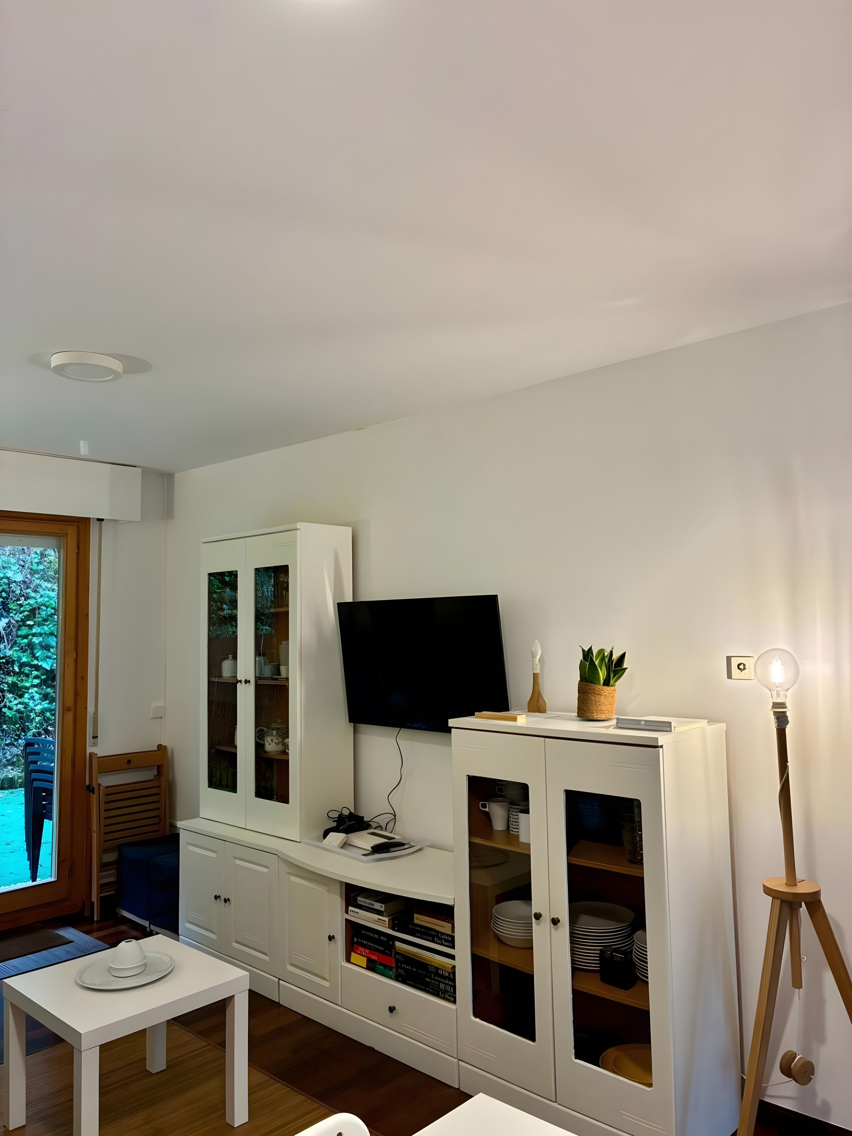 Una sala moderna con televisión de pantalla plana, muebles blancos, una mesa de comedor y una lámpara de pie.