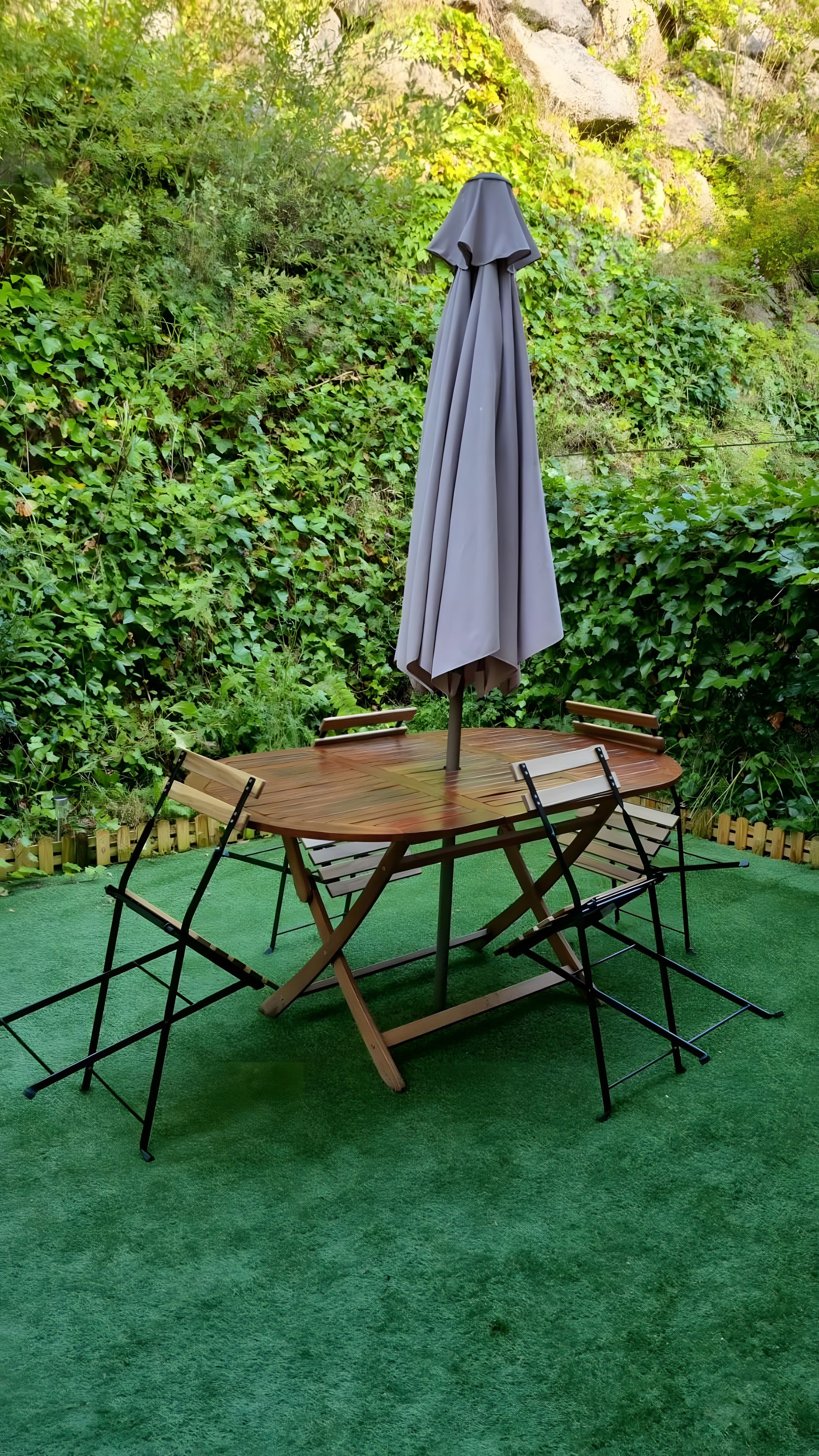 Una mesa de madera redonda con una sombrilla gris, rodeada por un seto verde.