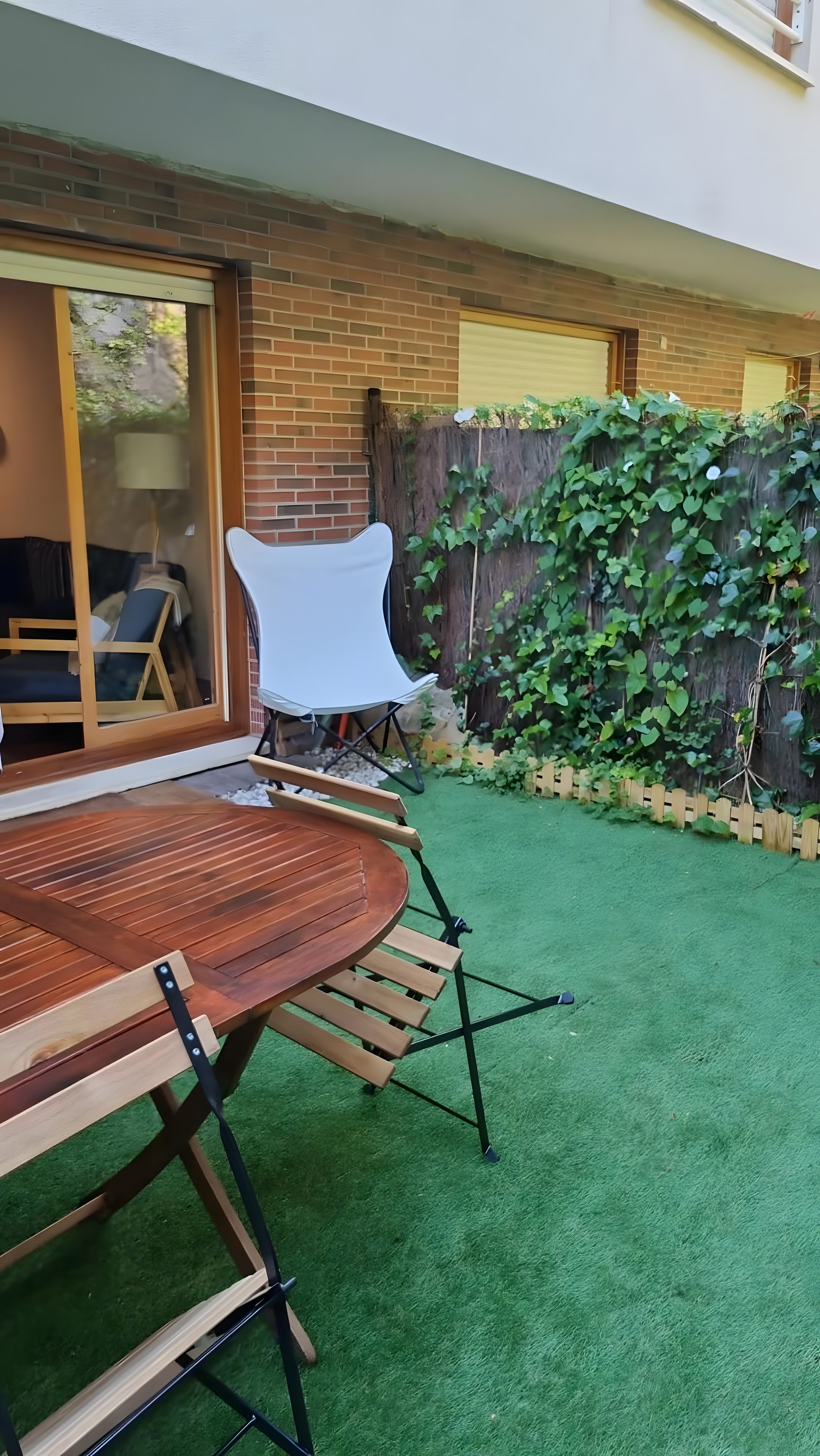 Un patio con una mesa y sillas de madera, una silla blanca y un muro de ladrillo con cerca de madera y follaje.