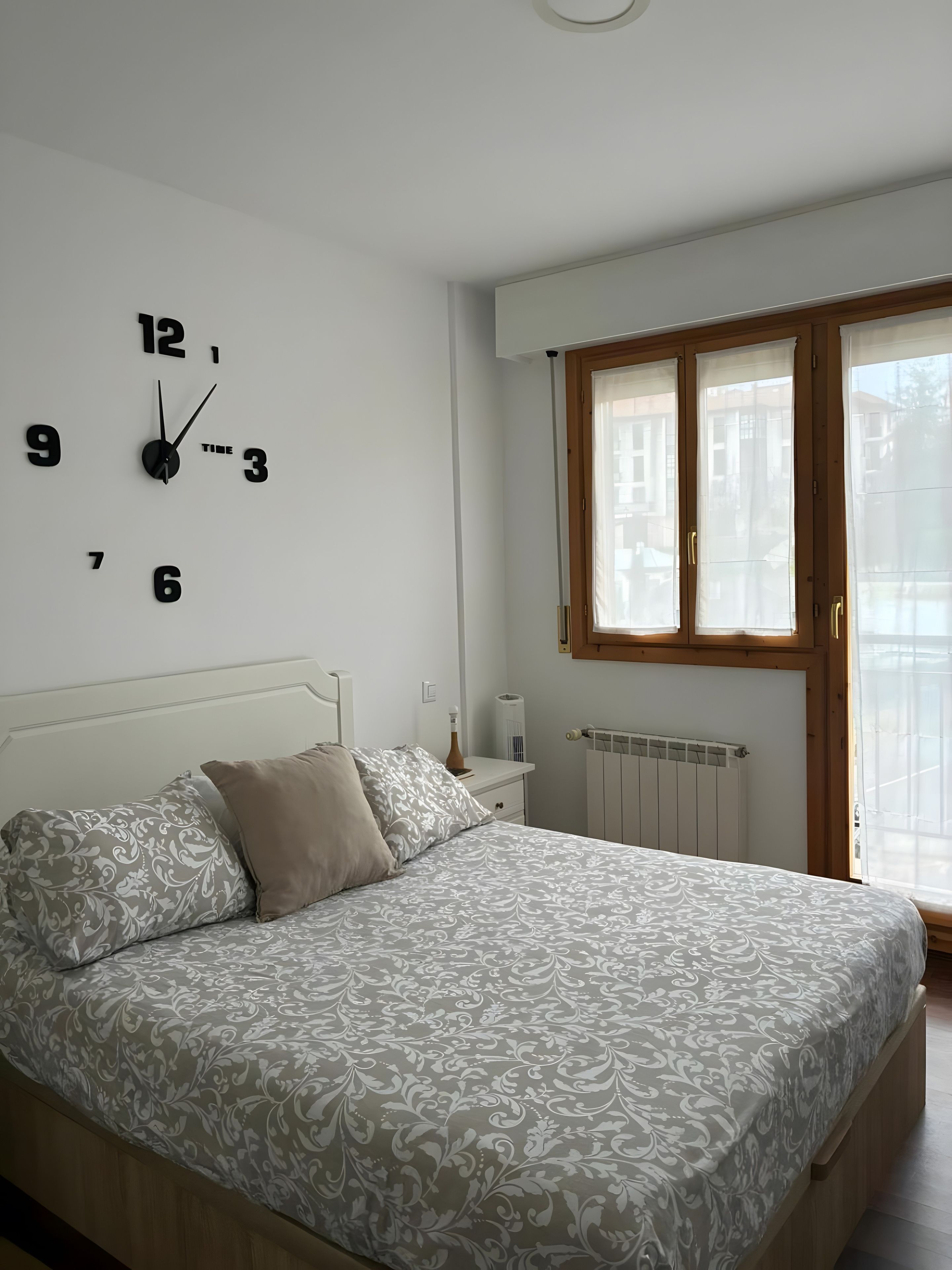 Un dormitorio con una cama, un reloj de pared, una ventana con cortinas y un radiador.