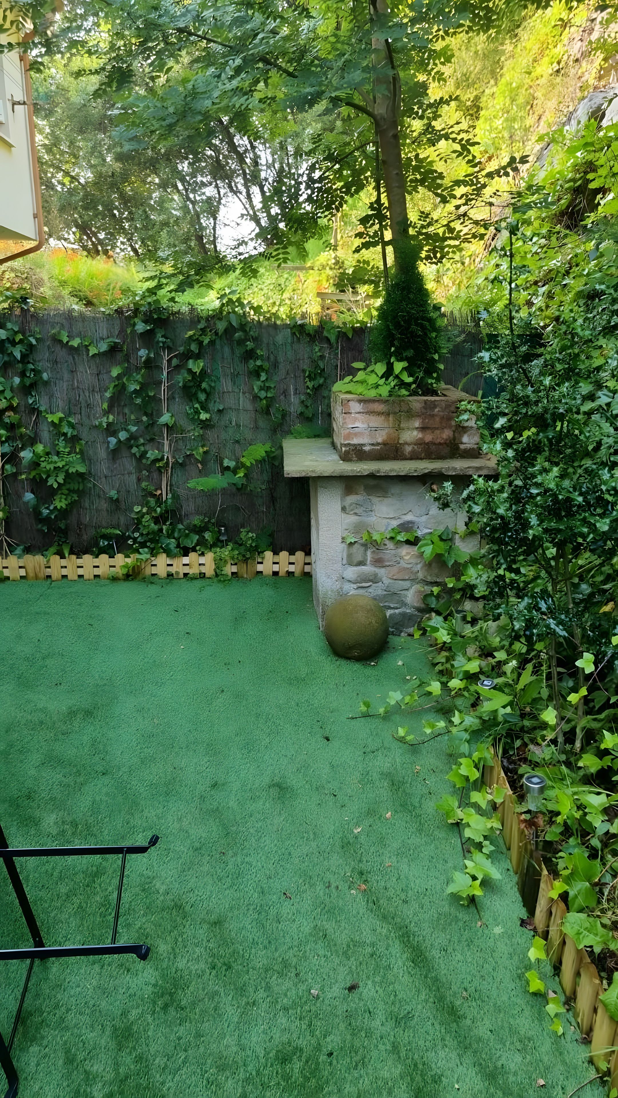 Un patio con césped artificial, cerca de madera, muro de piedra y caja de plantación de madera.