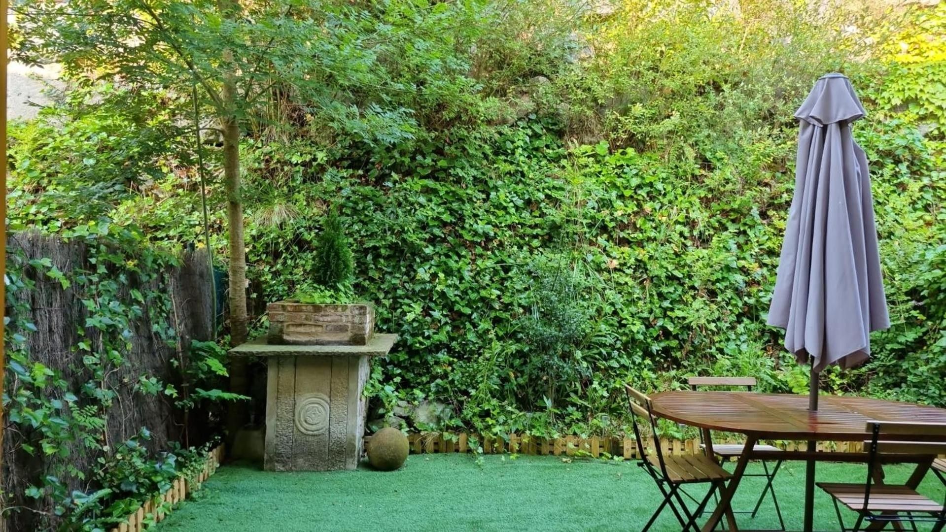 Un jardín con mesa y sillas de madera, un banco de piedra y un gran paraguas.
