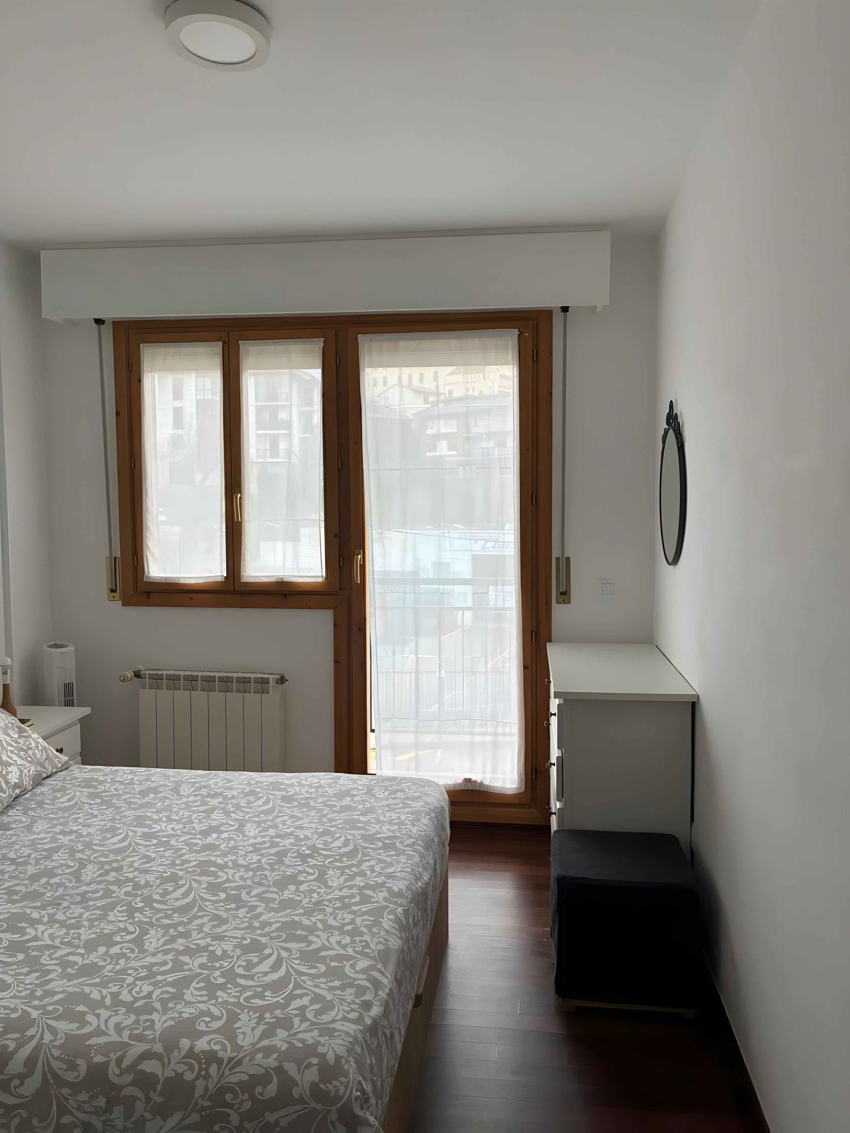 Un dormitorio con cama, un radiador, una ventana con cortinas, una mesita pequeña y un espejo en la pared.