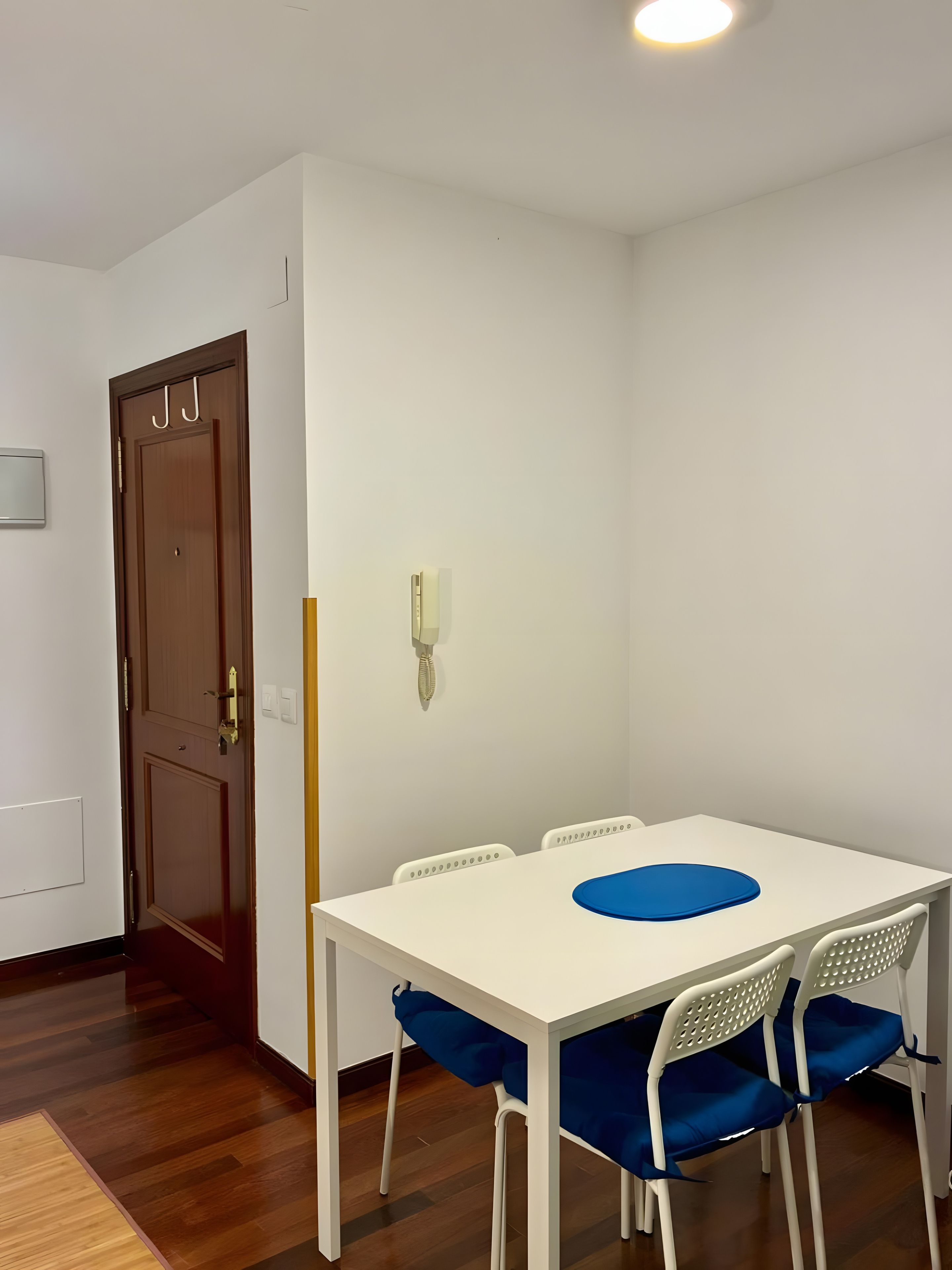Un comedor con mesa blanca y sillas azules, una puerta de madera y una pared blanca con un interruptor de luz.