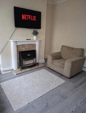 Een woonkamer met een open haard, een bank en een aan de muur bevestigde televisie waarop 'NETFLIX' wordt weergegeven.