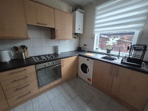 Een keuken met een gasfornuis, oven, wasmachine en een raam met uitzicht op een bakstenen muur en bloemen.