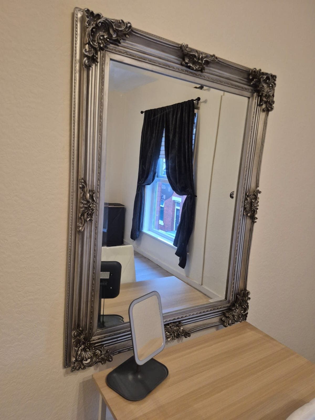 A mirror with an ornate frame on a wooden surface, reflecting a room with a window, curtains, and a chair.