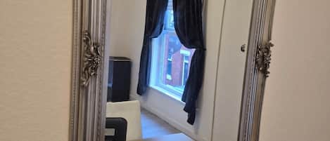 A mirror with an ornate frame on a wooden surface, reflecting a room with a window, curtains, and a chair.
