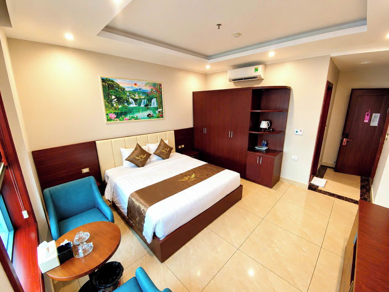 A hotel room with a bed, a blue armchair, a small table with a tray, a wardrobe, and a painting on the wall.