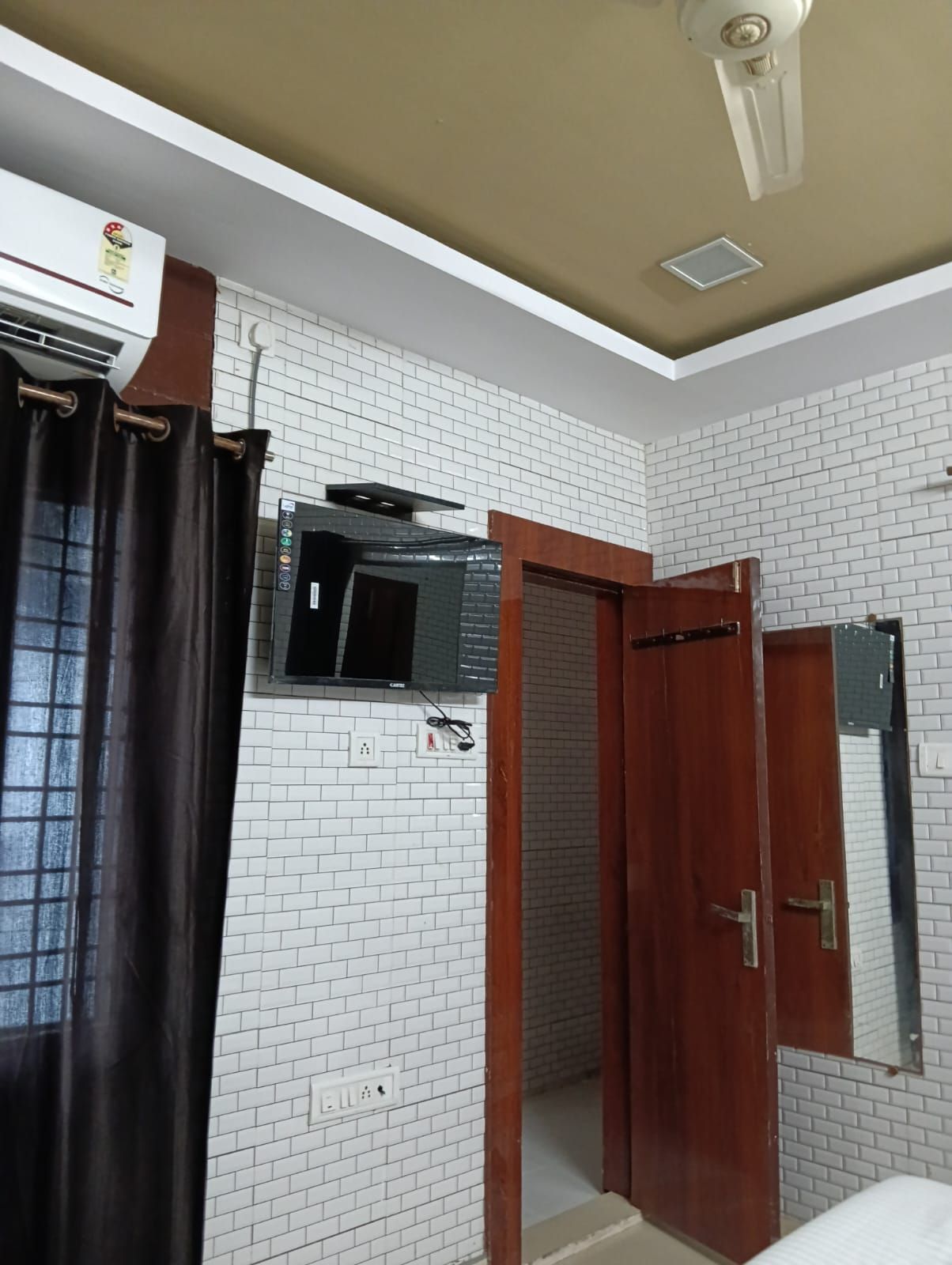 Una habitación con paredes de ladrillo blanco, un ventilador de techo, un equipo de aire acondicionado, una puerta de madera y una ventana con cortina.
