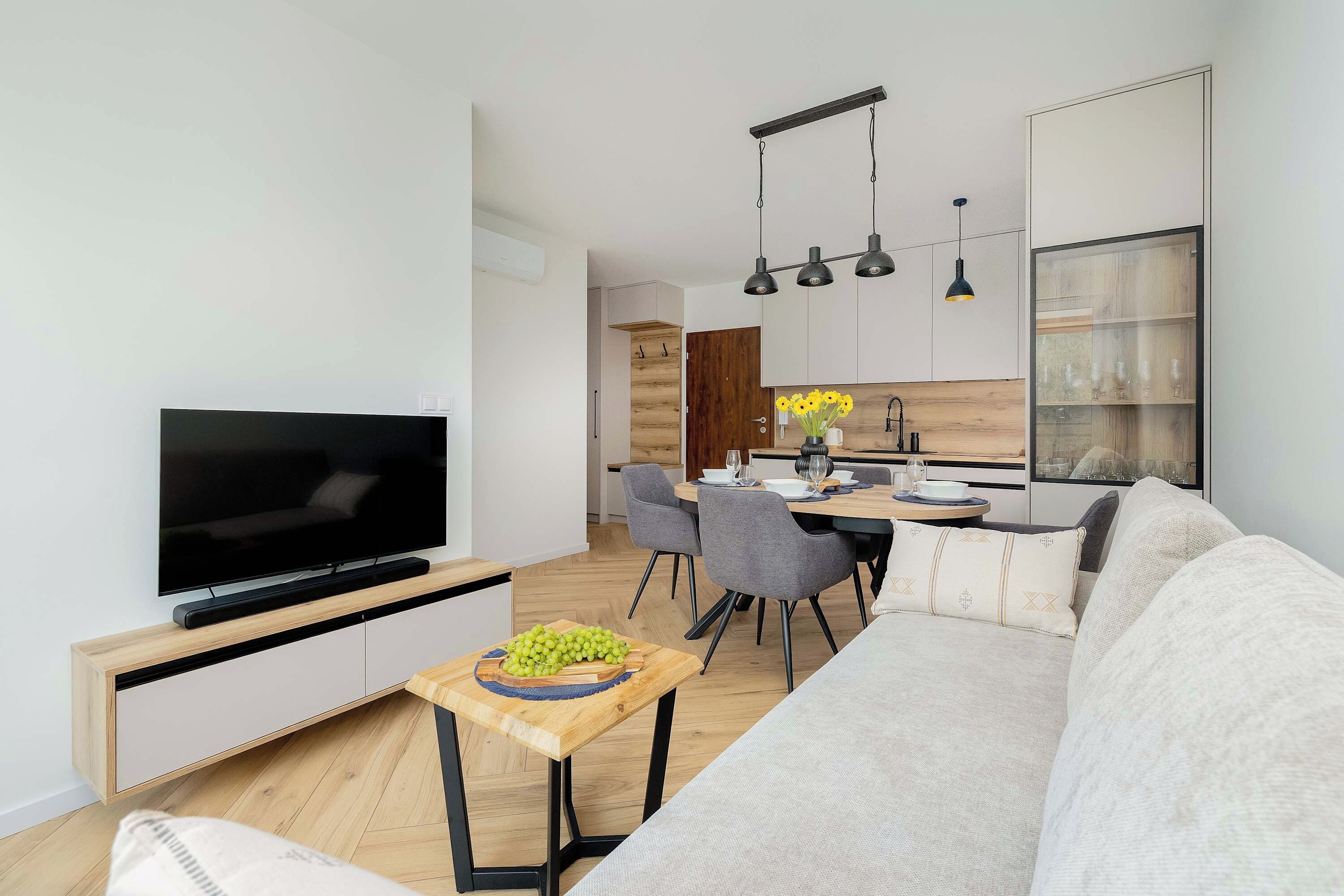 A modern living room with a flat-screen TV, a wooden coffee table with grapes, a grey sofa, and a dining area with a wooden table and chairs.