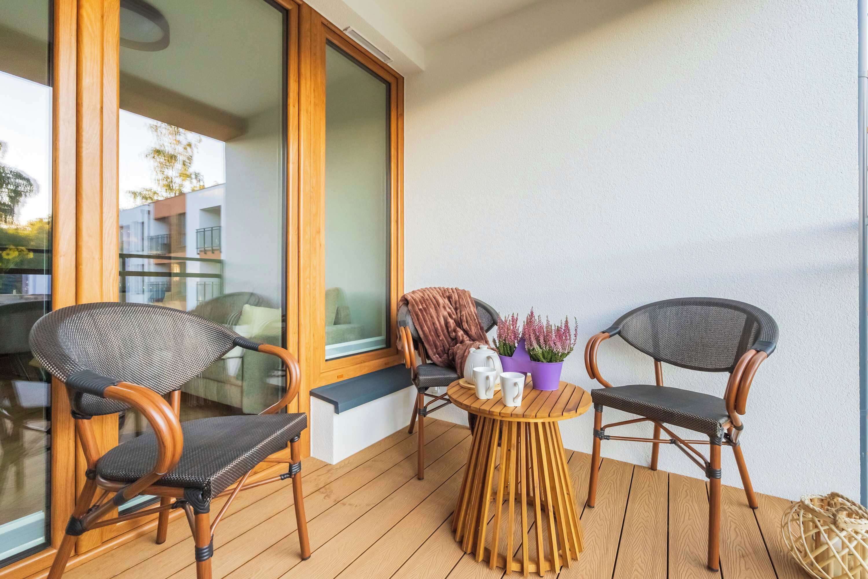 A balcony with two wicker chairs, a small round table with a vase of flowers, and a view of a building across the street.
