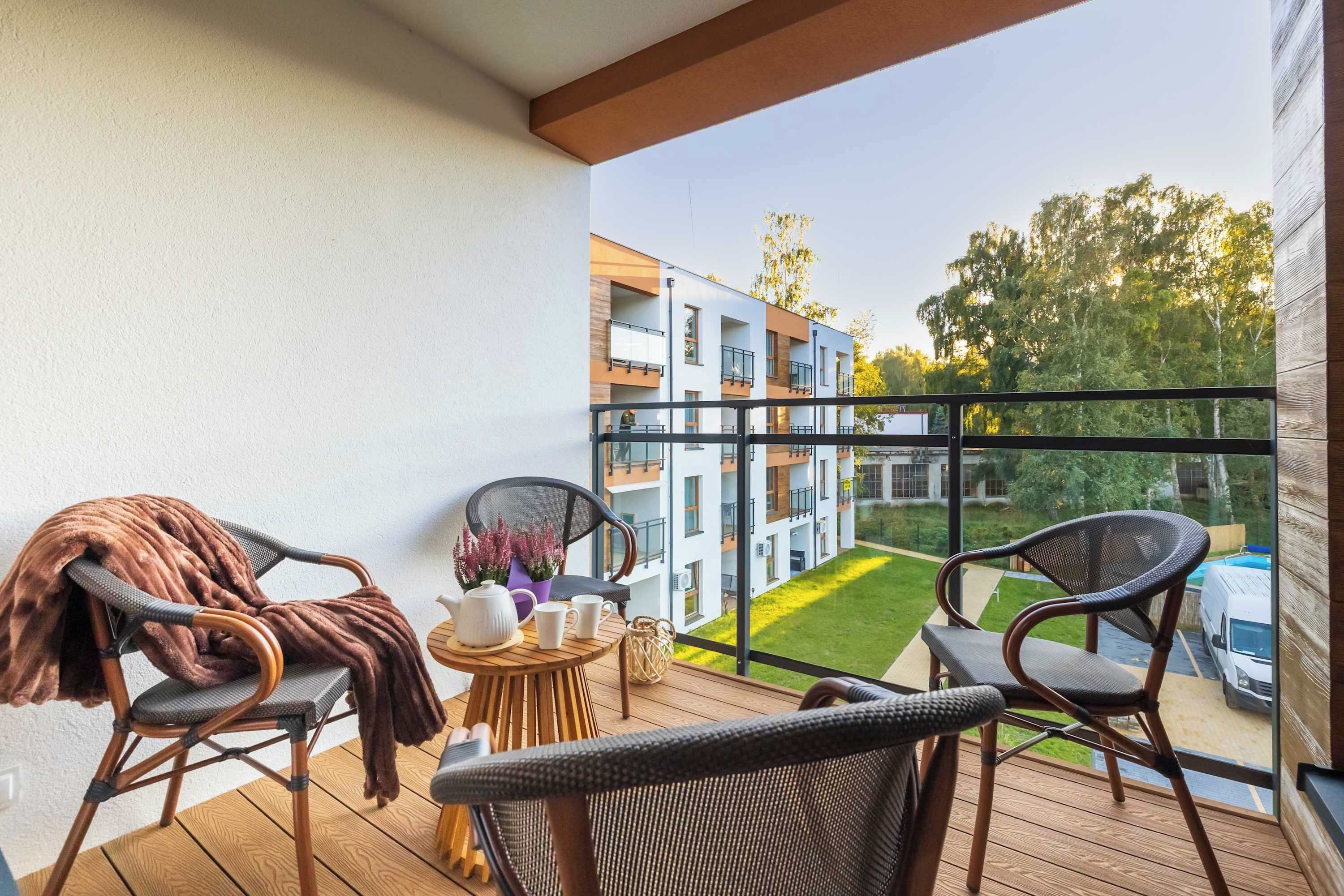 A balcony with wicker chairs, a small round table with a teapot and cups, and a view of a multi-story building with a green lawn.